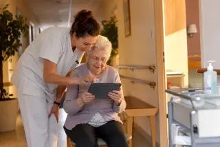 Personne âgée dans le couloir d’un EHPAD utilisant une tablette avec l’aide d’une infirmière, sous une lumière douce.