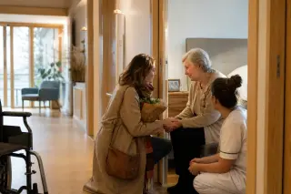 Femme adulte tenant la main de sa mère âgée dans un couloir d’EHPAD, avec une soignante à proximité, ambiance calme et lumineuse.