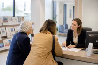 Personne âgée, un proche et un conseiller assis autour d’une table à la maison, examinant des documents non lisibles avec calculatrice et stylos, dans une lumière naturelle.