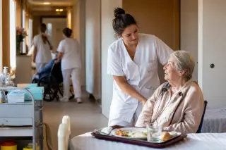 Aide-soignante accompagnant une personne âgée dans un couloir d’EHPAD, avec chariot de soins et lumière naturelle douce.