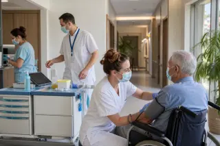 Infirmière en EHPAD prenant la tension d’un résident âgé dans un couloir lumineux, avec chariot de soins et aide-soignante à proximité.